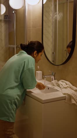 Confident-brunette-girl-with-Brown-skin-color-in-green-pajamas-is-working-at-the-sink-and-washing-her-hands-while-getting-ready-for-bed-in-the-evening-in-the-bathroom